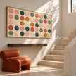 Wide Multicolor Painted Dot Grid Wall Art shown beside a staircase and an orange chair.